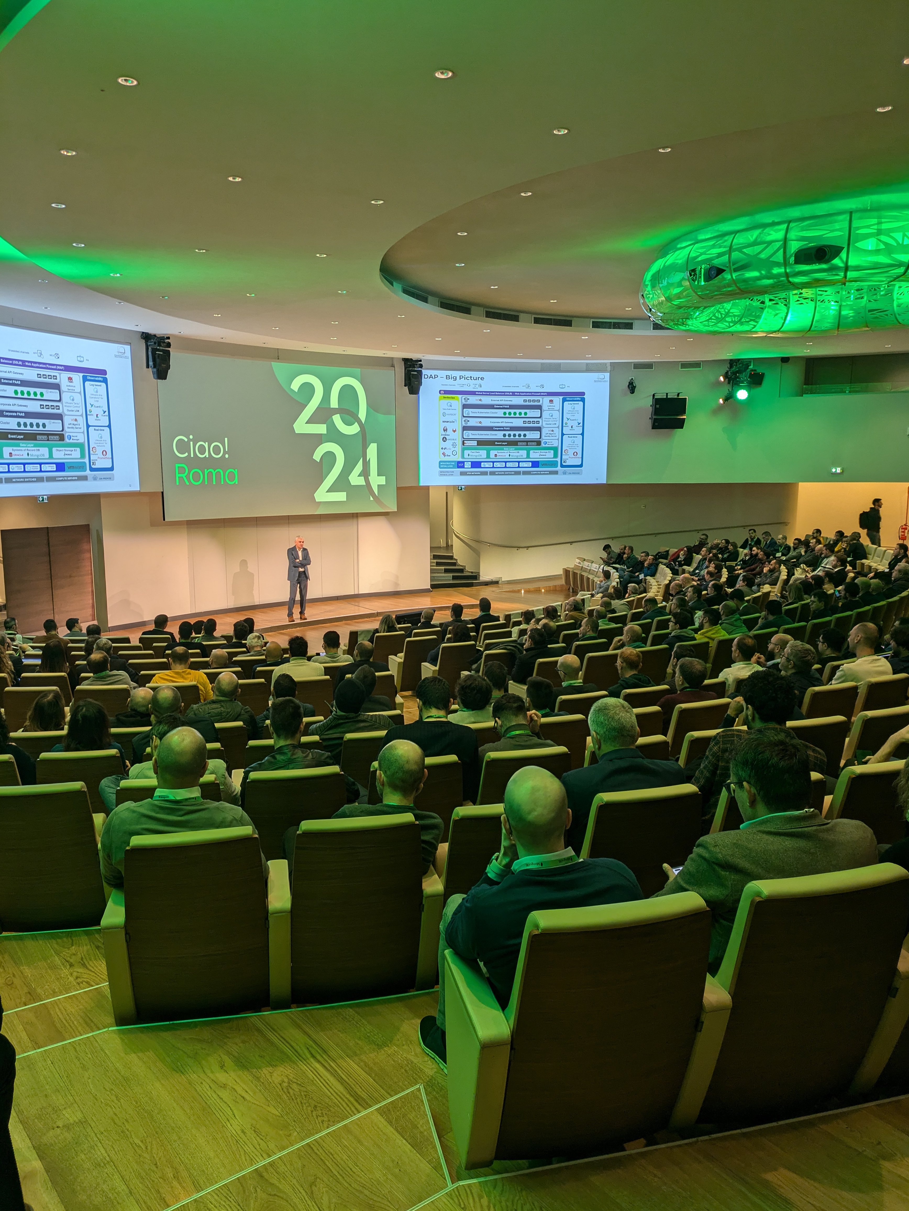 Sessione plenaria al MongoDB Day Roma 2024, con un relatore che parla sul palco davanti a una platea. Sullo schermo si legge 'Ciao! Roma 2024' con dettagli tecnici visibili nelle slide proiettate.